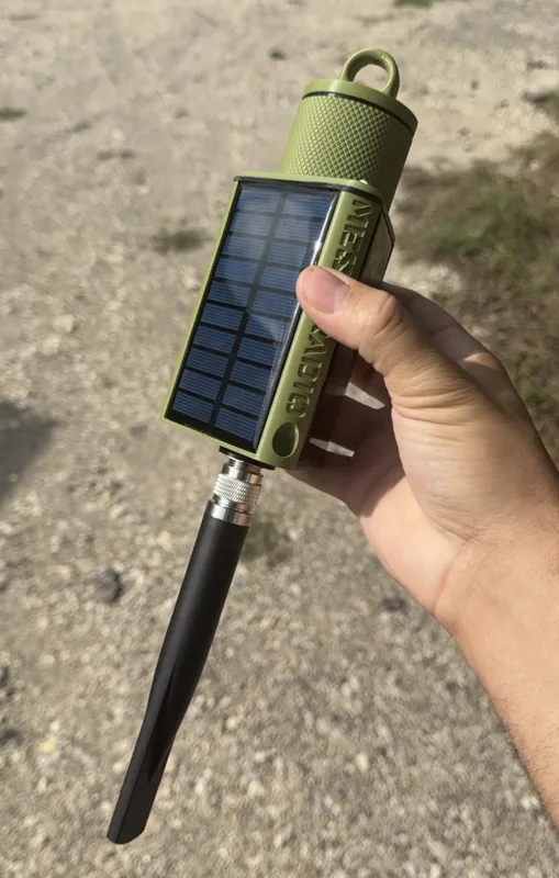 Olive green solar-powered Meshtastic radio with N-type antenna connector, built-in solar panel, carabiner clip, and ground spike